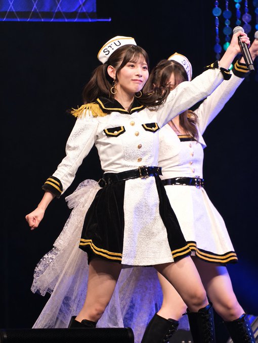 Two young women in matching white and black military-style idol costumes with gold trim and sailor hats perform on stage, one holding a microphone.