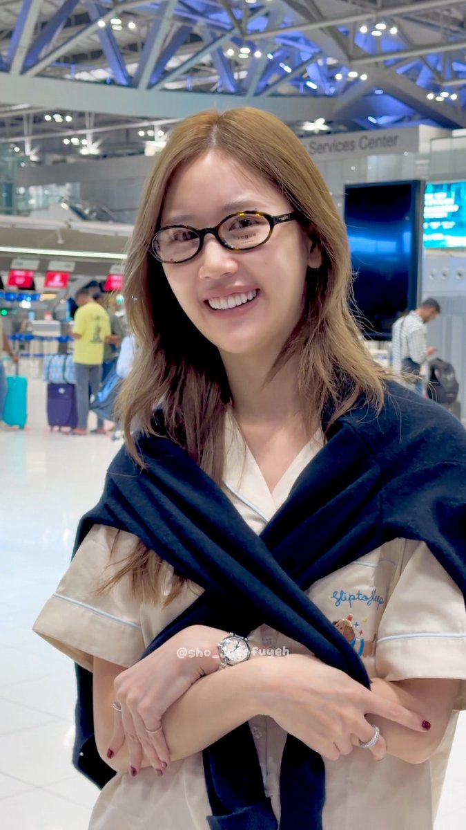 A smiling woman with long blonde hair and glasses poses at what appears to be an airport Services Center, wearing a cream top with a navy blue sweater draped over her shoulders.