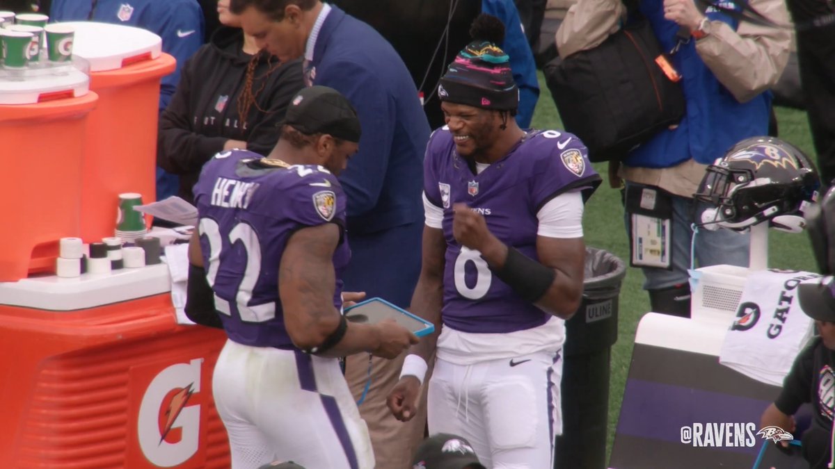Two Baltimore Ravens players in purple jerseys share a laugh on the sidelines, with #22 Henry facing #8 quarterback Lamar Jackson, who is smiling broadly while wearing a colorful pom-pom beanie and holding a tablet. The scene captures the fun, energetic sideline moment referenced in the post about Jackson being "mic'd up" and bringing energy, with a Gatorade cooler and Ravens helmet visible nearby.