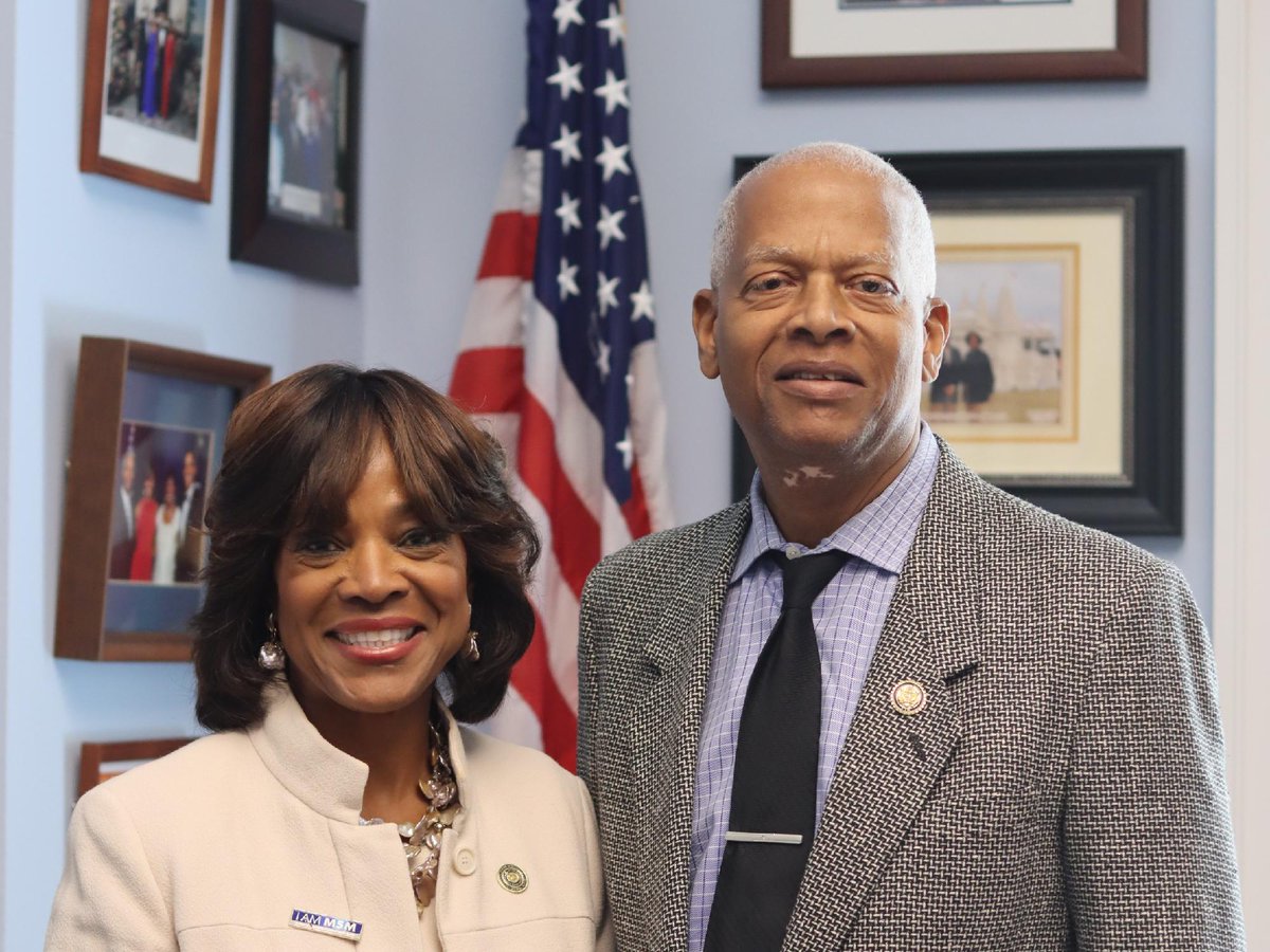 A woman and a man stand together facing the camera in an office. An American flag and framed photographs and artwork are visible behind them. There is text on the woman's jacket pin.