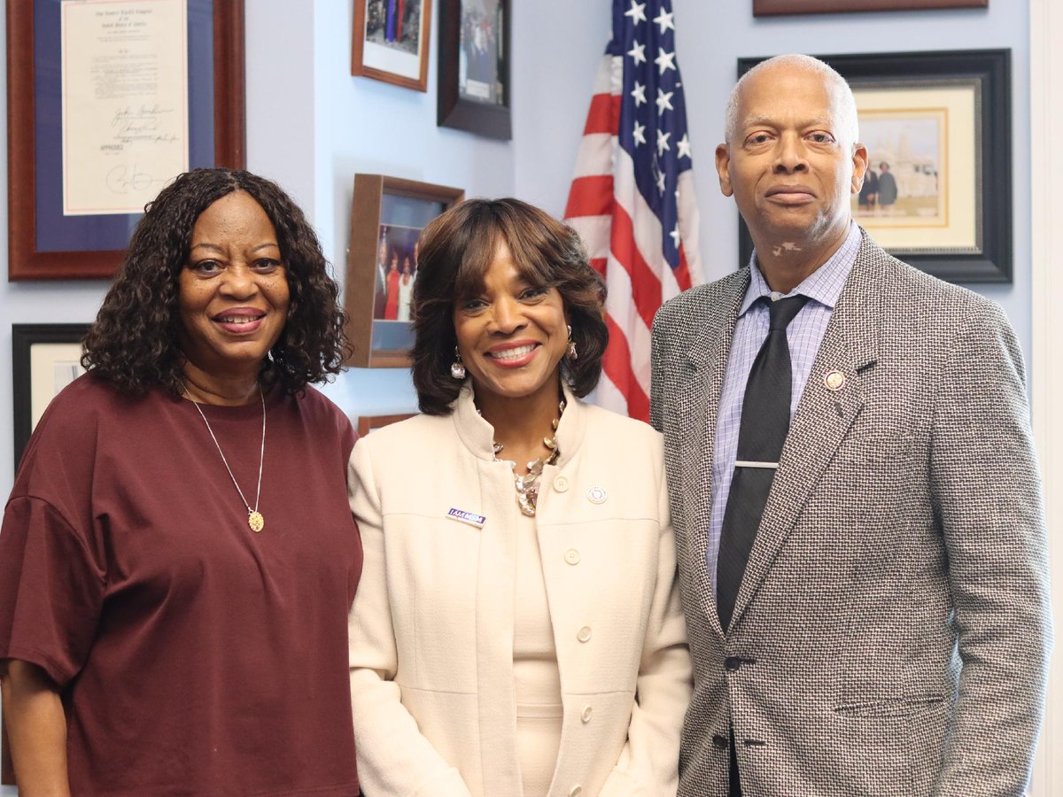 A posed photograph of three adults standing together in an office. Framed documents and photographs hang on the wall behind them, alongside an American flag. There is text on some of the framed items and clothing.