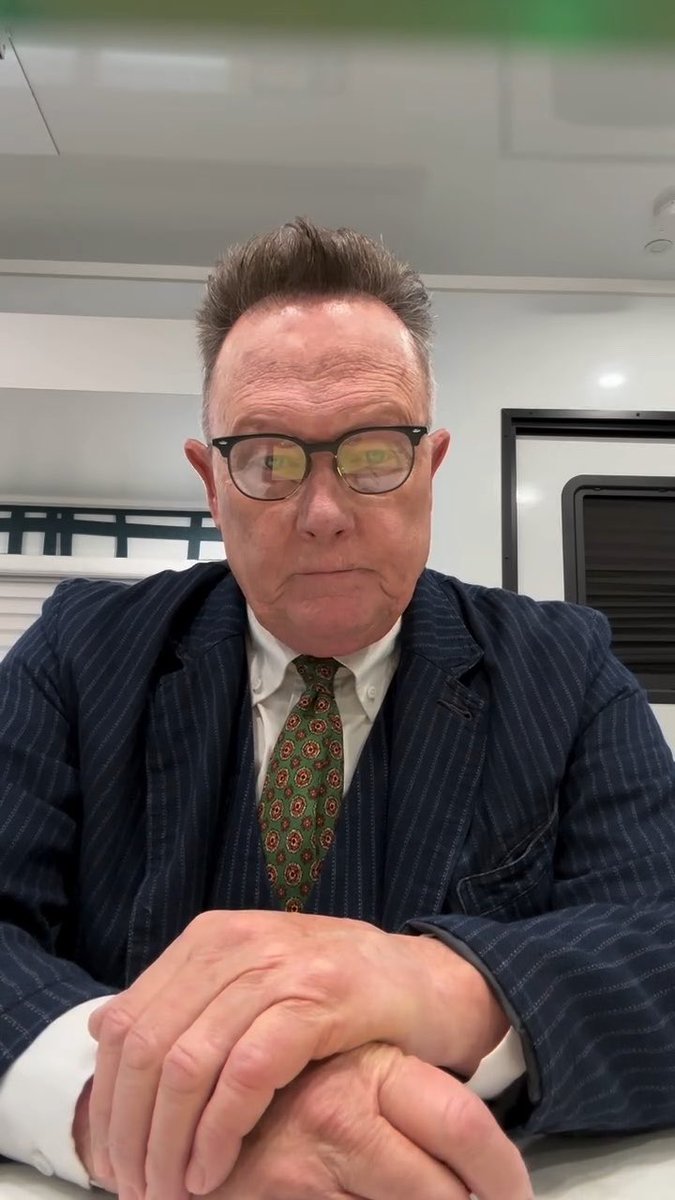 A close-up selfie of an older man wearing glasses, a striped suit jacket, white shirt, and green patterned tie, seated indoors.