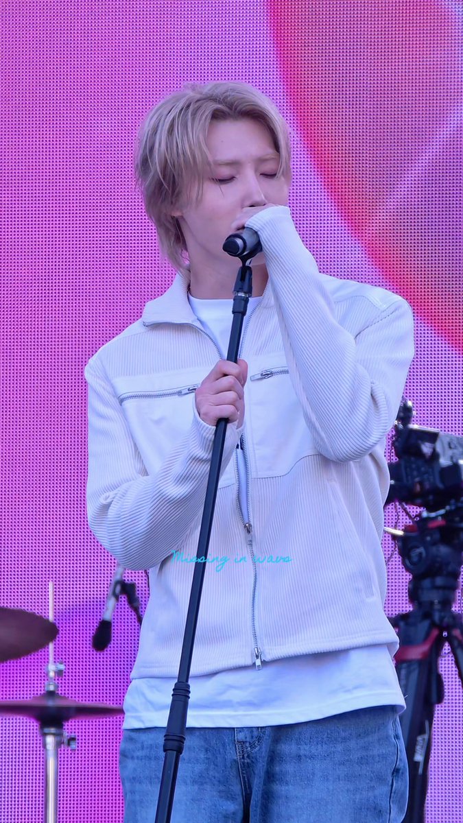 A person with short blonde hair sings into a microphone while wearing a white zip-up jacket, with eyes closed in concentration. A pink dotted backdrop is visible behind them, along with a watermark reading 'Morning in mars'.