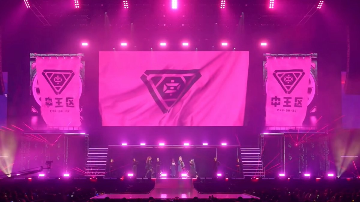 A concert stage bathed in pink and red lighting, featuring large screens displaying a triangular logo with the text '中王区' and 'CHU-OU-KU', with performers visible on the stage below.