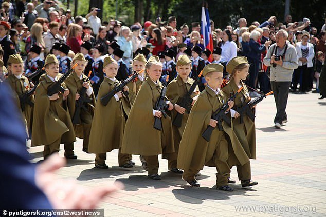 A parade scene featuring children in long olive-green military capes marching in formation with rifles, watermarked 'pyatigorsk.org/east2west news'.