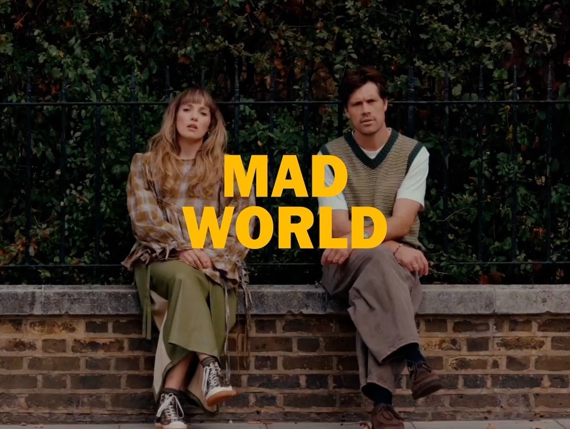 A man and woman in stylish autumn attire sit side by side on a brick wall with greenery behind them, with the bold yellow text "MAD WORLD." displayed prominently across the center of the image, reinforcing the post's caption through its prominent visual title treatment.