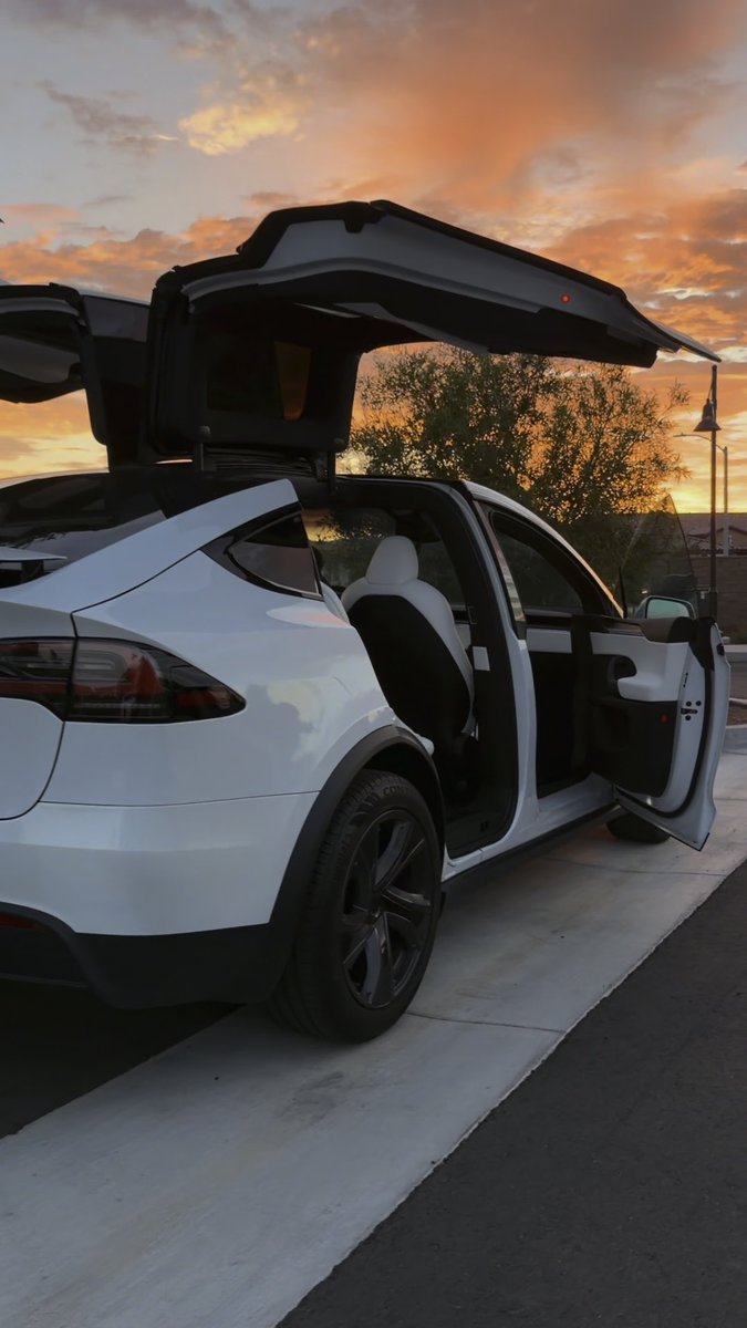 A white Tesla SUV is parked with its falcon-wing rear doors and passenger doors open against a backdrop of a vibrant orange sunset.