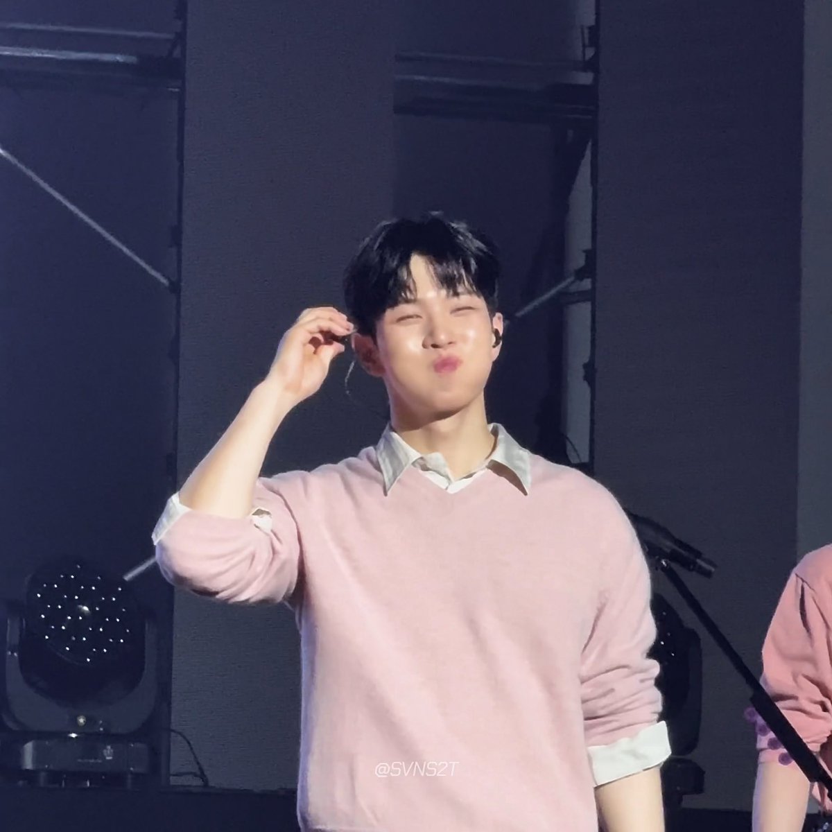 A young man in a pink sweater over a white collared shirt makes a kissing face on stage, adjusting an in-ear monitor.