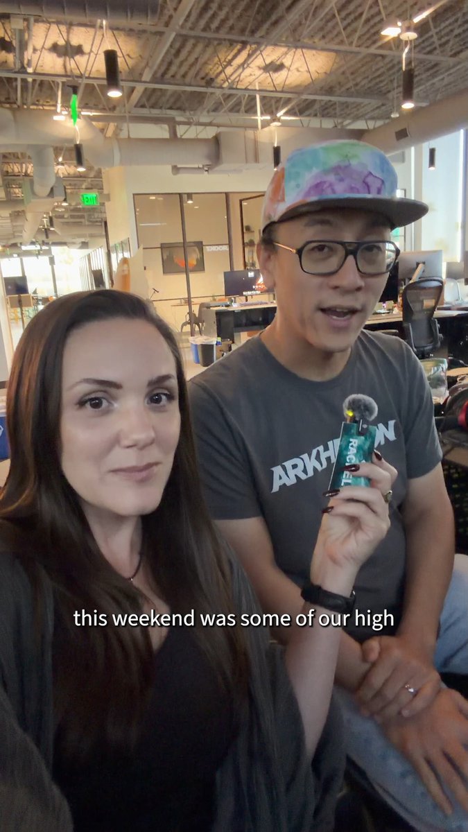 A man and woman pose together indoors in an open workspace, with the man holding a small microphone device and the subtitle 'this weekend was some of our high' visible.