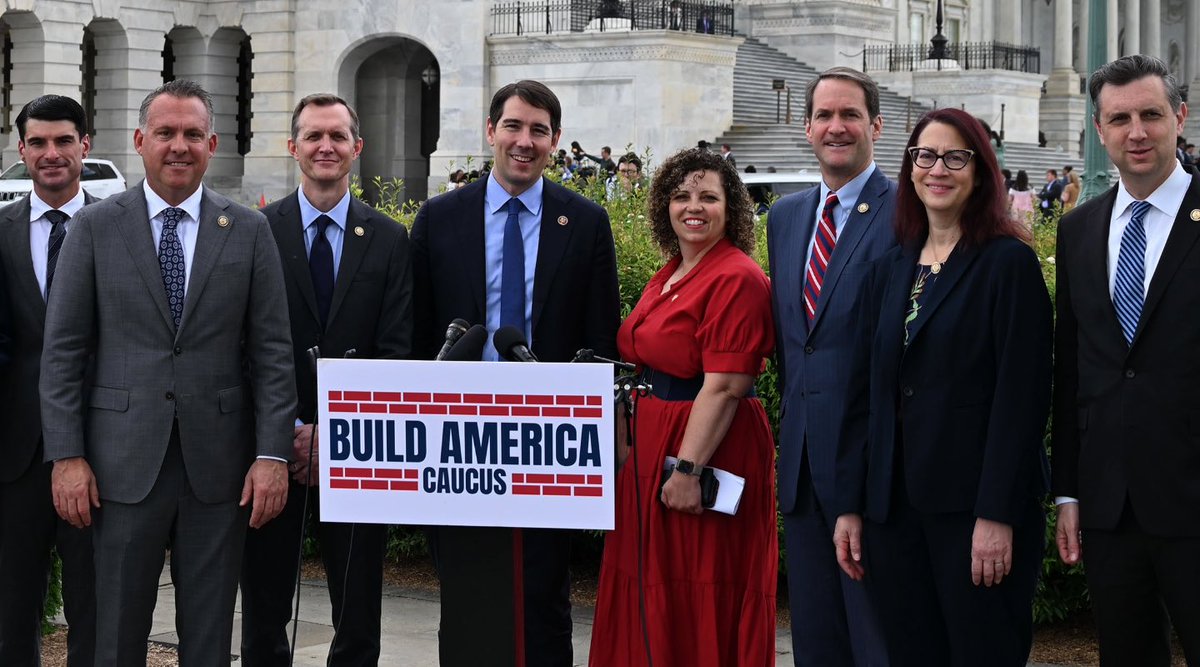 A group of formally dressed people pose outdoors in front of a government-style stone building. One person stands behind a podium with microphones and a sign; plants, steps, and railings are visible in the background.