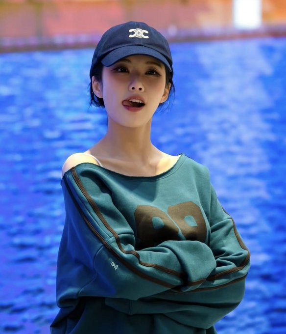 A young woman wearing a navy blue Chanel baseball cap and a teal/green sweatshirt with the number '69' on it, posed in front of a blue textured background.