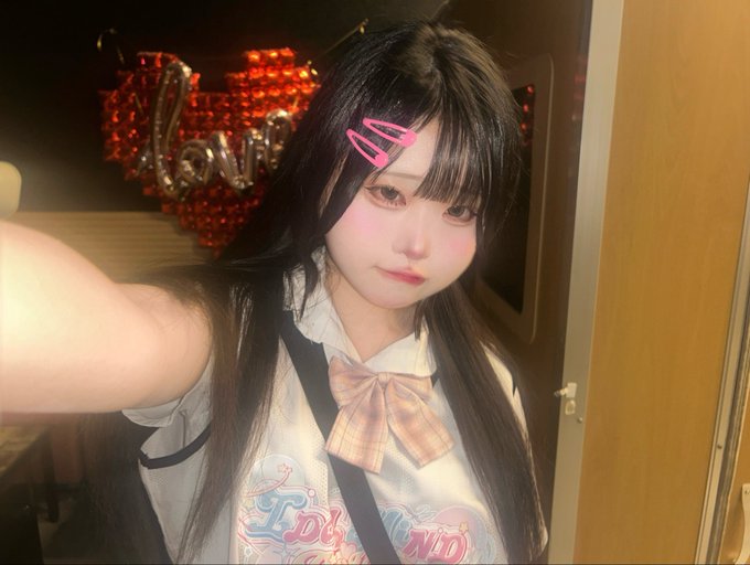 A selfie of a young woman with long dark hair and pink hair clips, wearing a white ruffled shirt and plaid bow, with a red heart balloon in the background.