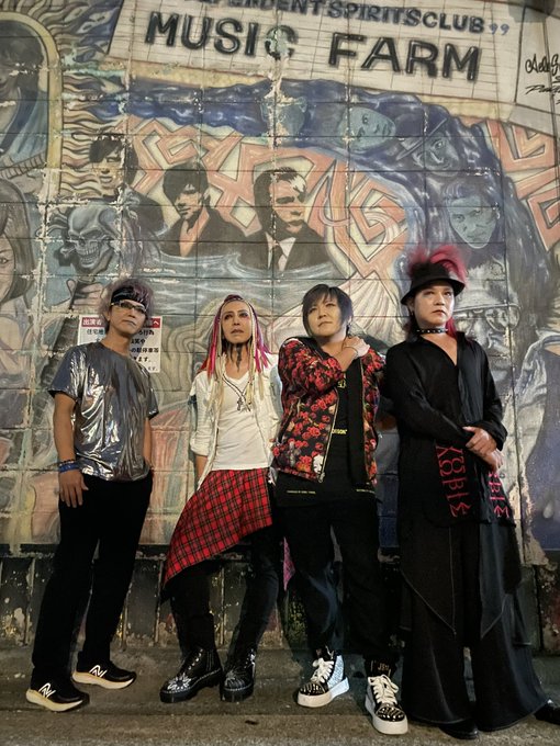 Four people in alternative fashion pose in front of a wall featuring a mural of musicians and text reading "MUSIC FARM" and "STUDENT SPIRITS CLUB."
