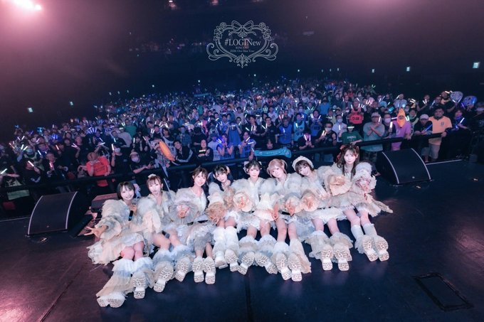 A large group of performers in matching white frilly outfits with cream-colored stuffed accessories sitting in a row on stage, facing a packed audience at a venue. A logo reading '#LOGNew' is displayed on the screen above the crowd.
