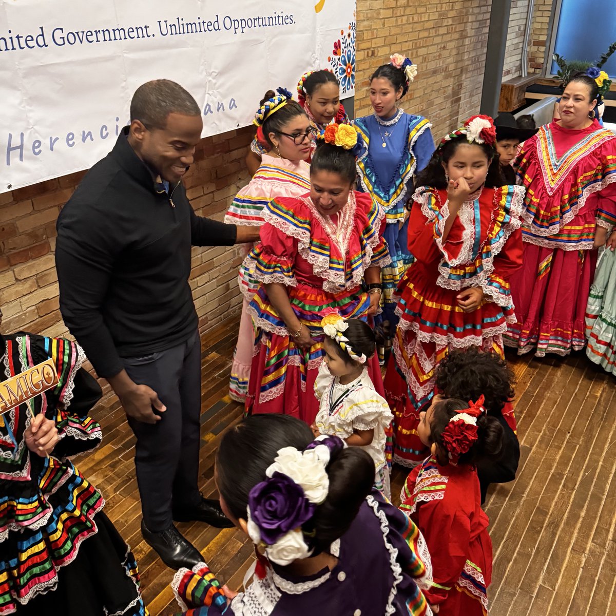 A man in dark clothing stands with a group of women and children wearing colorful traditional dresses and floral hair accessories, gathered indoors beside a brick wall and a banner. There is text on the banner.