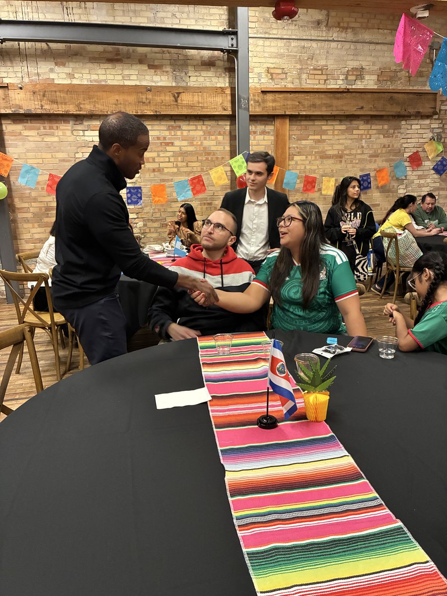 A man in a black outfit shakes hands with a seated woman wearing a green soccer jersey at a decorated indoor gathering. A seated man in a red hoodie looks on, with other people gathered around tables in the background. The foreground table has a colorful striped runner, a small flag, a potted plant, and drinking glasses. There is some visible text.