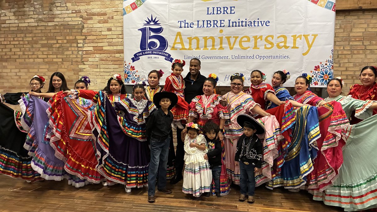 A group of adults and children pose together in colorful traditional dance costumes in front of a large anniversary banner. There is text on the banner.