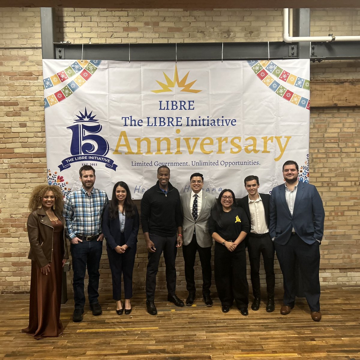 A group of eight adults pose for a photo in front of a colorful anniversary banner hanging on a brick wall.