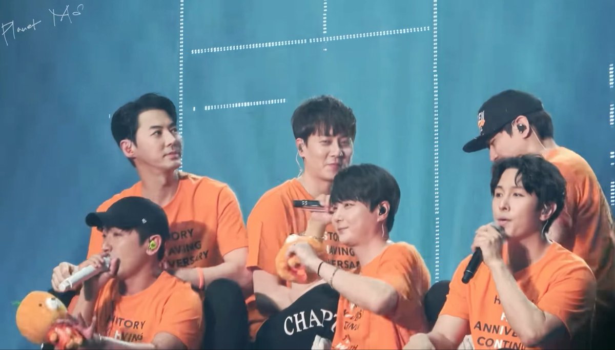 Six members of a K-pop group in matching orange "History" anniversary t-shirts perform on stage, holding microphones with smiles.