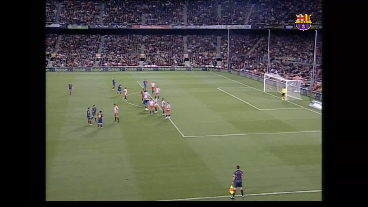 A packed stadium scene shows players positioned near the penalty area as a player in a blue and red striped jersey (FC Barcelona, per the logo) readies for a free kick, with a goalkeeper in yellow set near the goalpost and an assistant referee holding a flag visible in the foreground—capturing the moment of Leo Messi's first career free-kick goal.