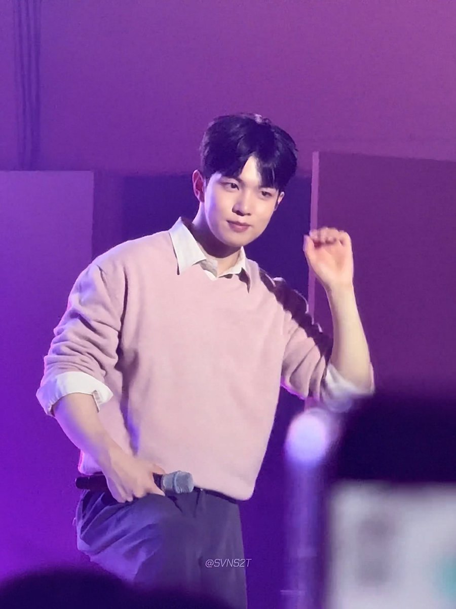 A young man in a pink sweater holds a microphone on stage under purple lighting, with the watermark '@SVN52T' visible.