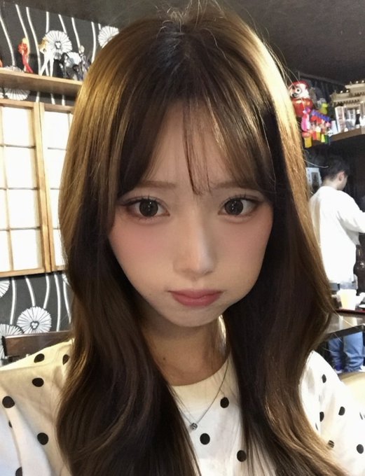 A close-up selfie of a young woman with long brown hair and bangs, wearing a white top with black polka dots and a silver necklace.