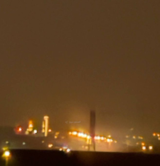 A blurry nighttime photograph of a city skyline illuminated by orange lights, with Arabic text watermark 'المصور أونر' across the image.