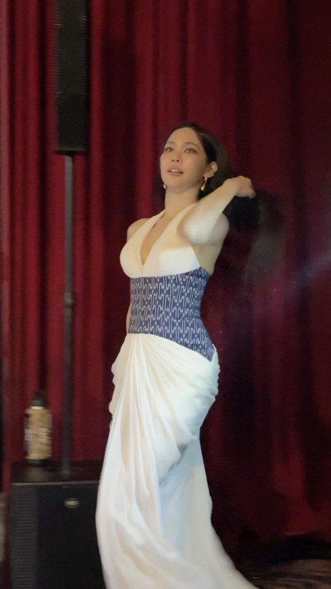 A woman with dark hair wearing a white halter dress with a blue patterned corset, dancing or posing in front of a dark red curtain.