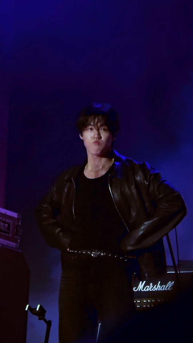 A man with dark hair wearing a black leather jacket over a black shirt, standing against a purple-lit background with his hands on his hips; 'Marshall' is visible as a watermark.