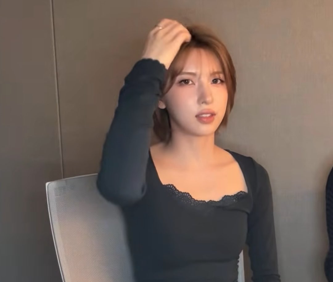 A woman with brown hair wearing a black long-sleeved top with a square neckline and lace detail sits in a chair, raising one hand toward her head against a beige wall background.
