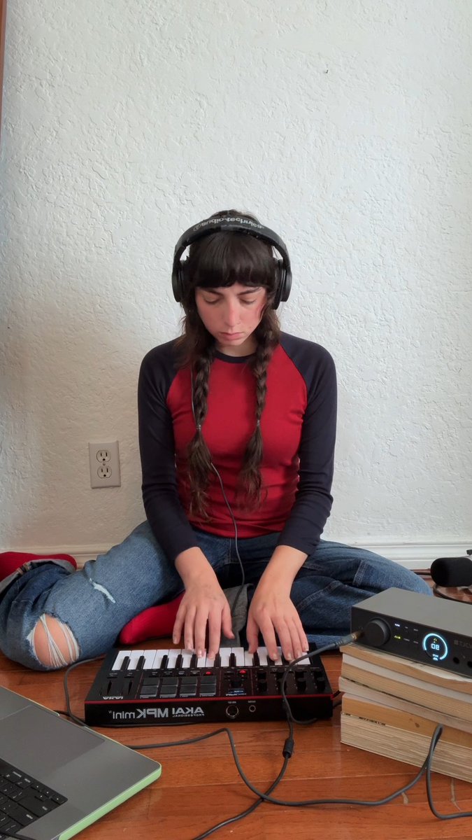 A person with dark braided hair, wearing headphones and a red and black long-sleeved shirt, sits cross-legged on the floor playing an "AKAI MPK Mini" MIDI controller.