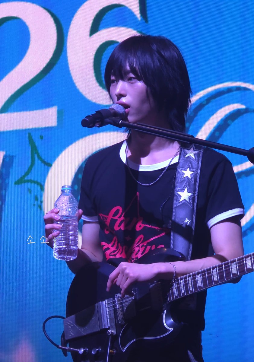 A performer with short black hair singing into a microphone while holding a water bottle and playing a black electric guitar on stage against a blue backdrop.