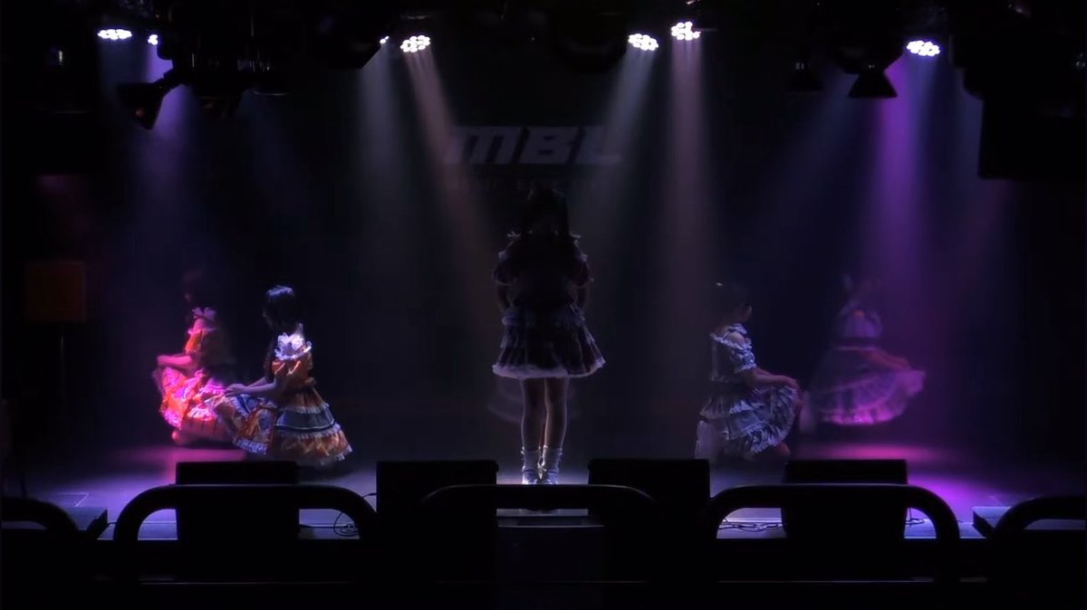 A dimly lit stage performance featuring dancers in colorful frilly costumes under purple spotlights, with partial "MBL" text on the backdrop.