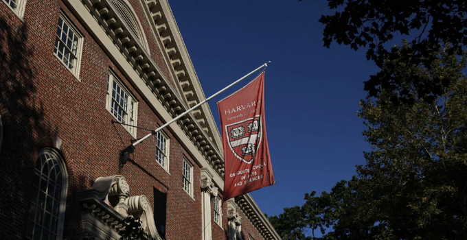 A red Harvard banner reading 'HARVARD UNIVERSITY' and 'VERITAS' hangs from a brick building against a deep blue sky.