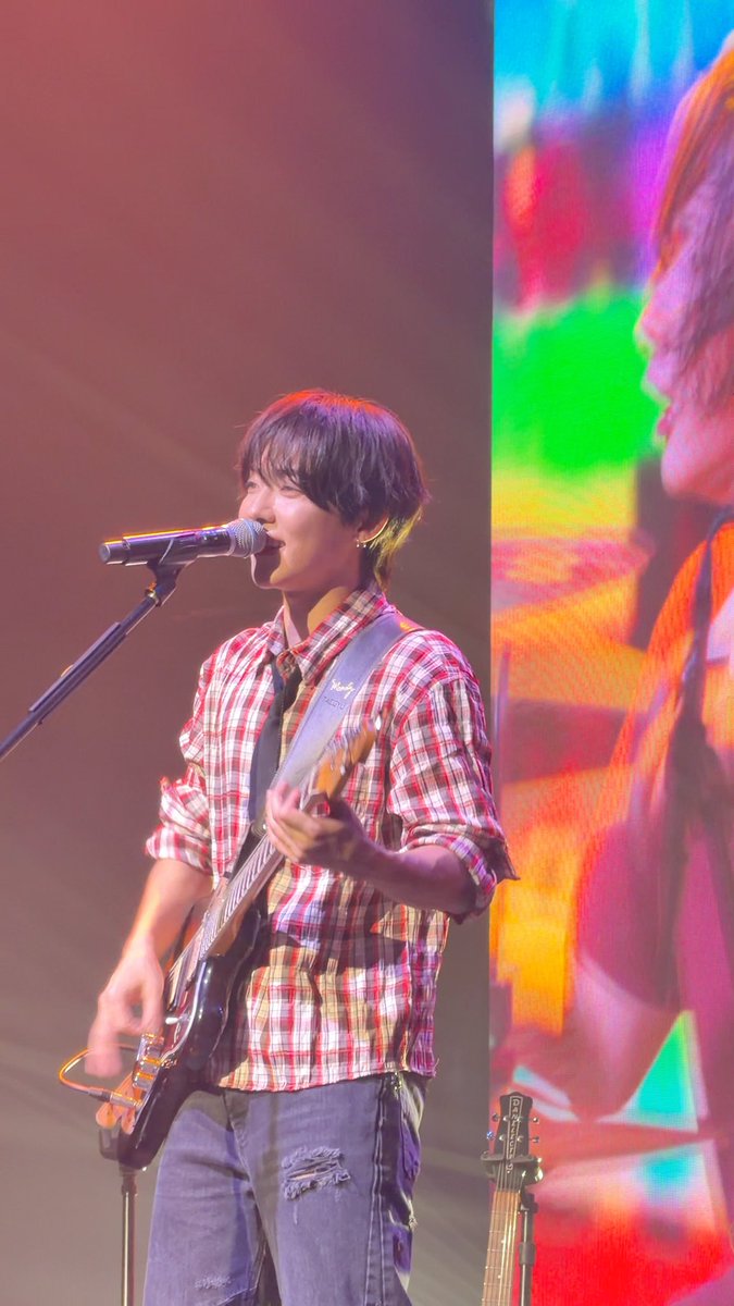Photo of a male musician with dark hair performing on stage, singing into a microphone while playing an electric guitar.