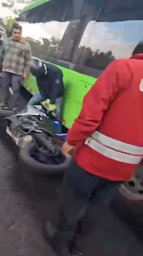 A motorcyclist wearing a helmet kneels beside a damaged bike with a broken wheel while a man in a red jacket and another bystander assist at the scene of a traffic collision. The image relates to the post documenting a multiple-vehicle accident at Periférico Sur and Camino a Santa Teresa, as captured on video.