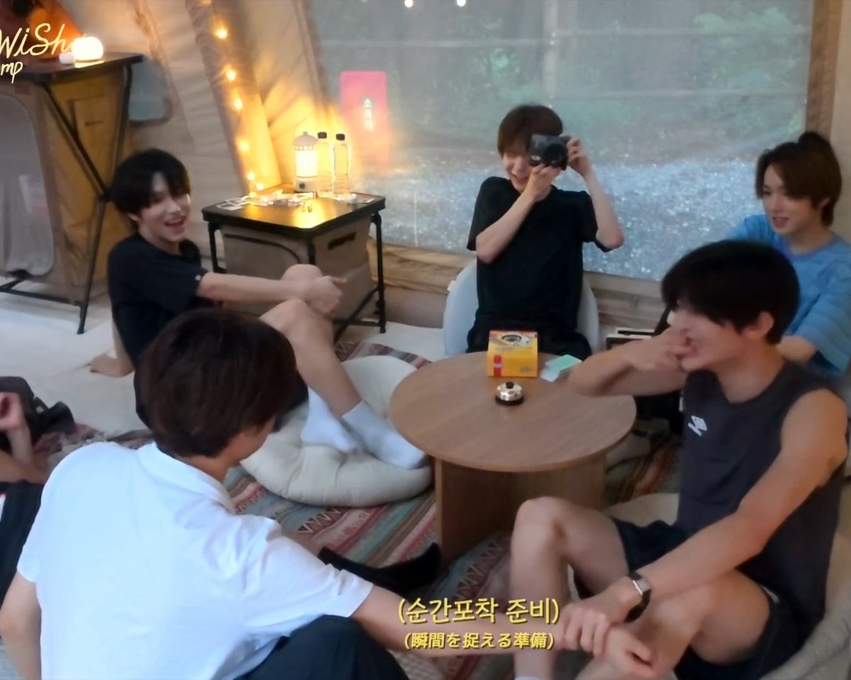Five young men sit in a tent-like setting with string lights, one holding a camera, with Japanese subtitles reading '(순간포착 준비)' and '(瞬間を捉える準備)'.