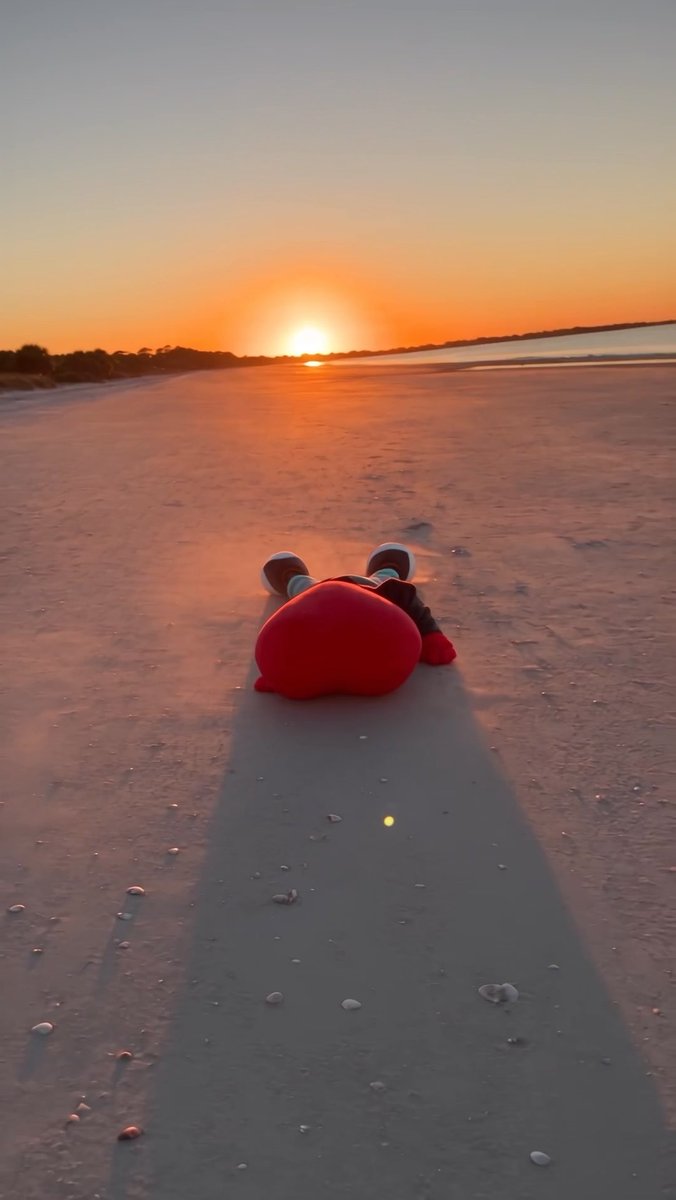 A red heart-shaped plush toy lying on a sandy beach at sunset, with the sun low on the horizon casting warm orange light.