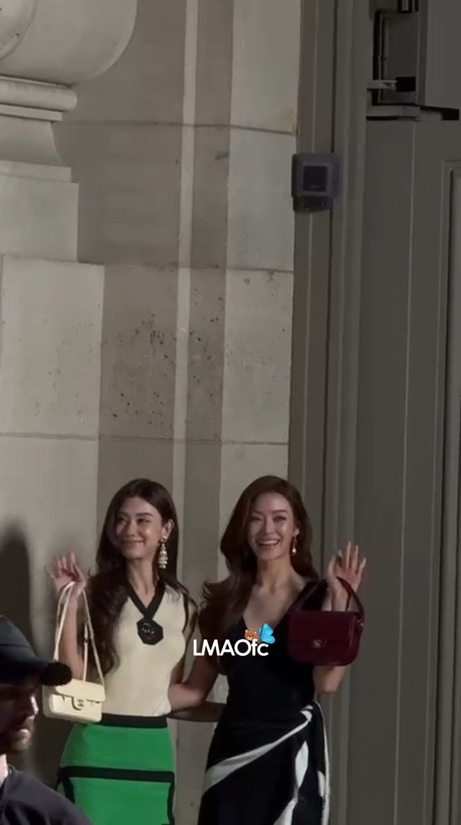 Two smiling women wave while walking together at night, one carrying a white handbag and the other a dark red one; a watermark reads 'LMAOfc'.