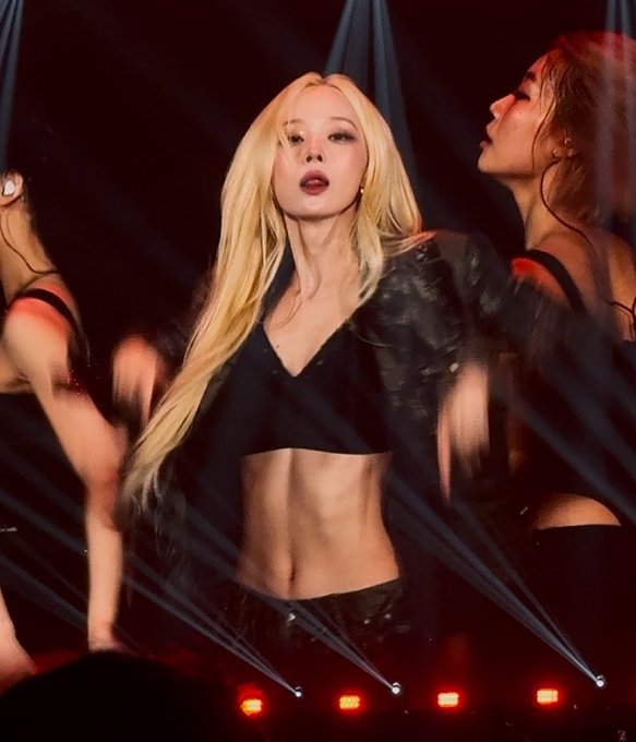 Image 10: A blonde performer in a black crop top and jacket stands center stage, flanked by backup dancers in black, with red stage lights and laser effects illuminating the performance.
