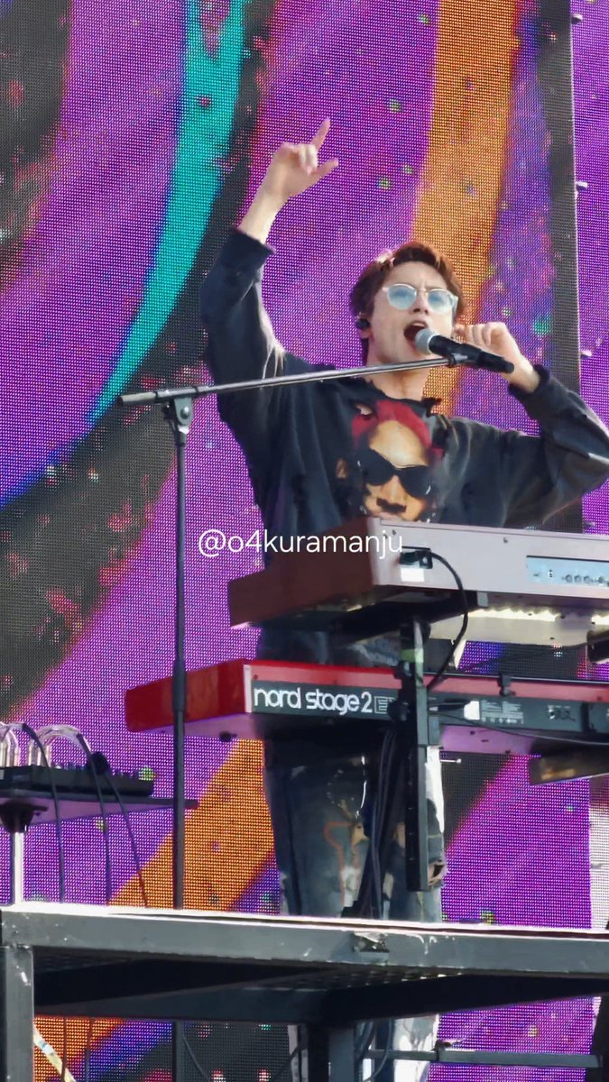A musician wearing glasses and a black jacket performs on stage, singing into a microphone with one hand raised while playing a Nord Stage 2 keyboard.