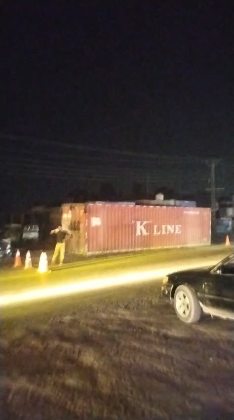 A nighttime scene shows a pink shipping container marked "K LINE" with a person standing nearby and traffic cones, with a dark car in the foreground.