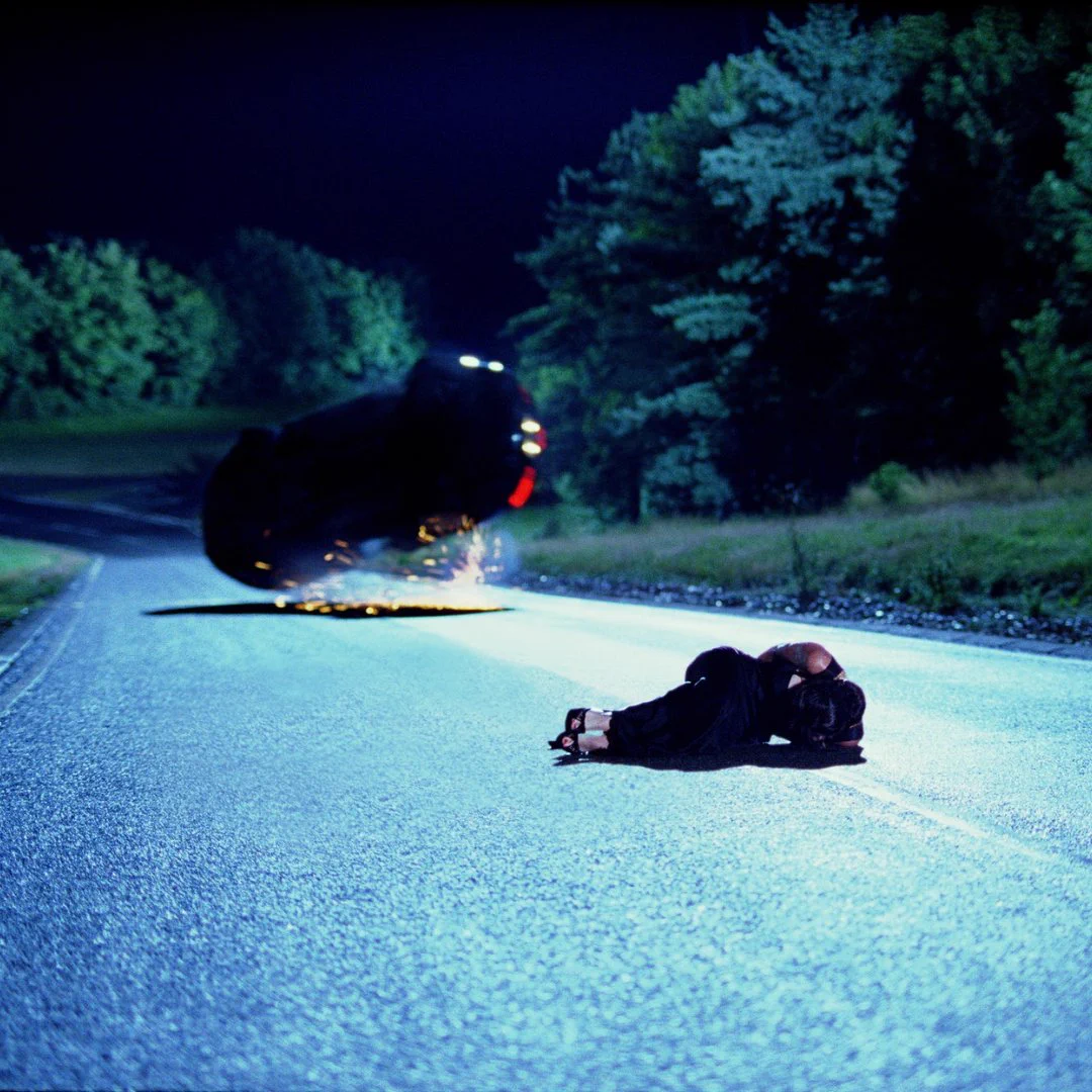 An image from Casino Royale. Vesper (Eva Green) is lying in the road as Bond (Daniel Craig) is driving in an Aston Martin DBS. The car has flipped and is rolling in the background.