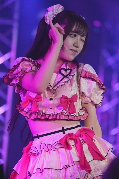 An idol with long brown hair and a high ponytail wearing an elaborate pink and white frilly outfit performs on stage, posing with her hand near her face.