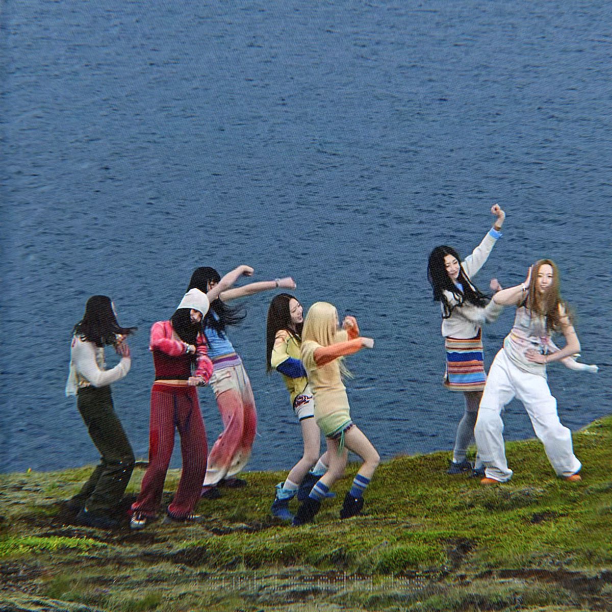 A group of young women dance energetically on a grassy hillside near a body of water.