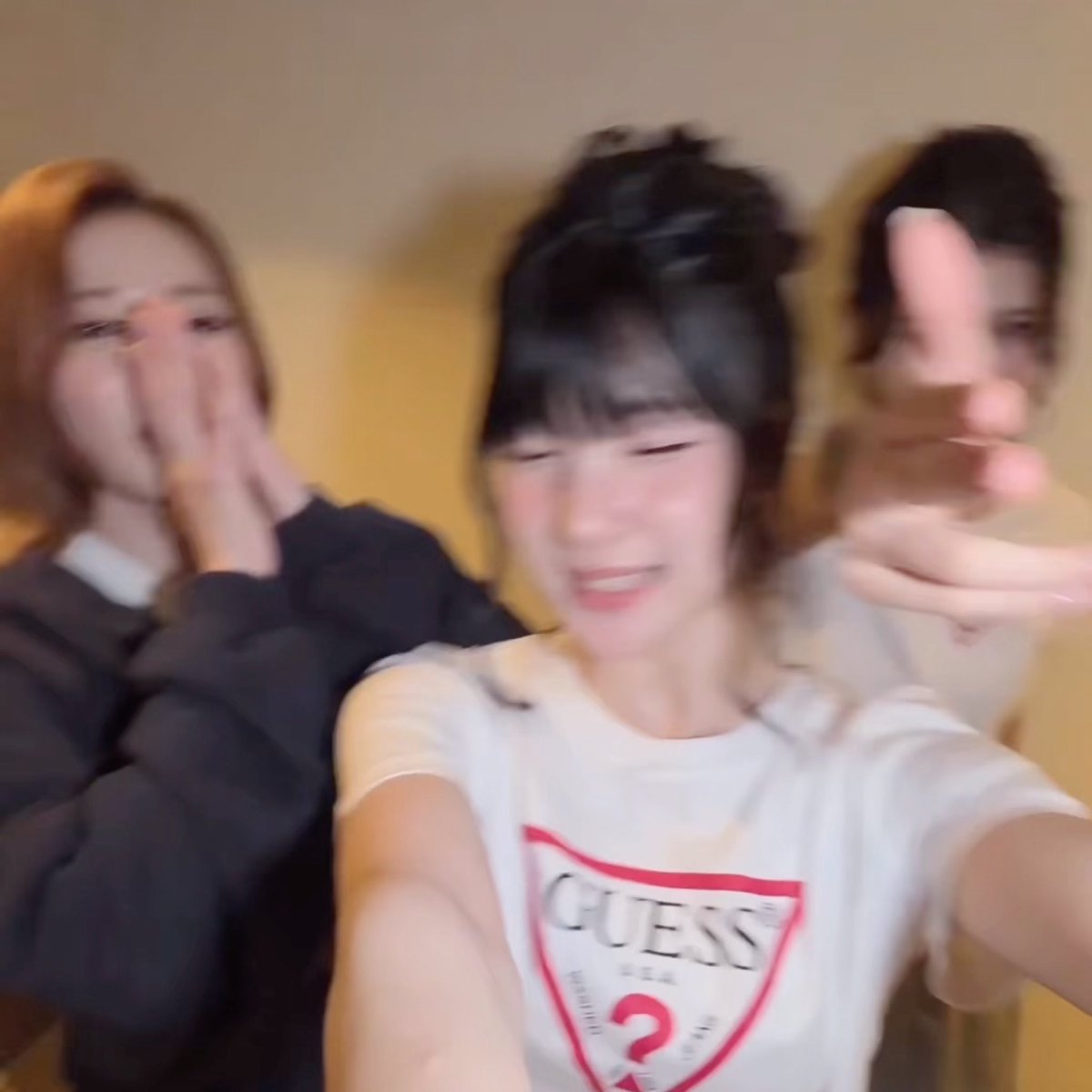 A blurry selfie of three young women, with the central figure wearing a white Guess t-shirt smiling while her friends pose behind her.