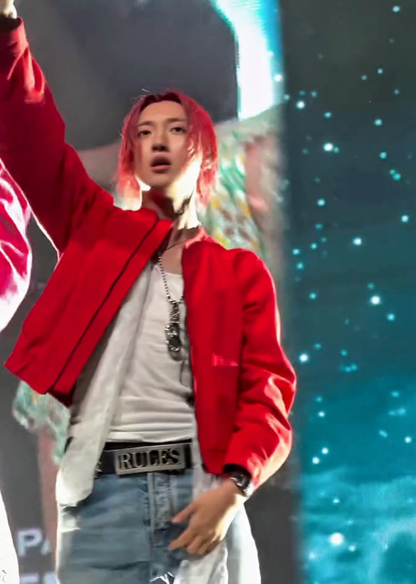 Woojin performs in Cologne wearing a vibrant red bomber jacket over a white t-shirt, paired with light blue jeans featuring a prominent "RULES" belt, while striking a dramatic pose with one arm raised against a sparkling teal stage backdrop.