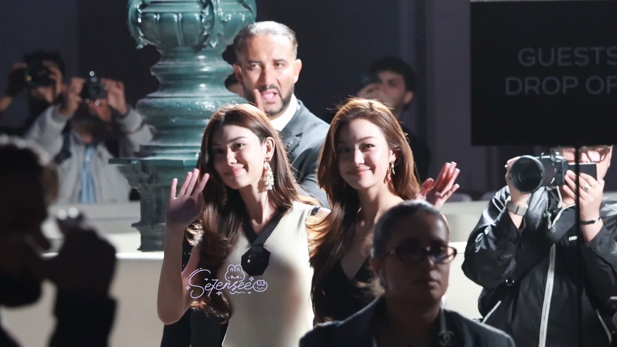 Two young women wave to the camera at what appears to be an event, with photographers and a sign reading "GUESTS DROP OF..." in the background. A watermark with the text "Seerisee" and cartoon graphics is overlaid on the image.