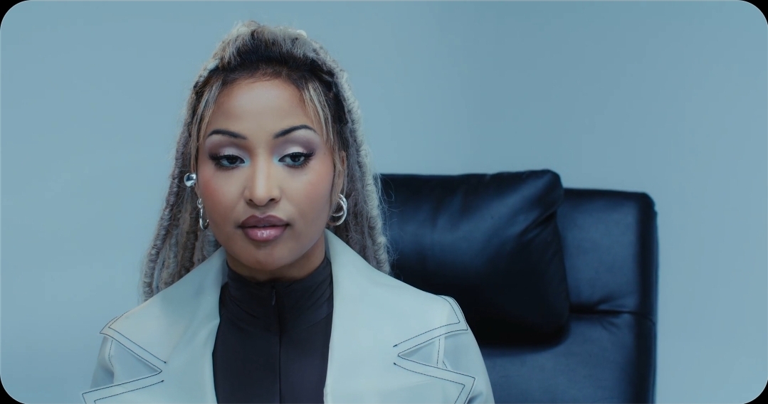 A portrait-style photo of a woman with blonde highlighted hair, wearing a white blazer over a black top, seated in a black office chair.