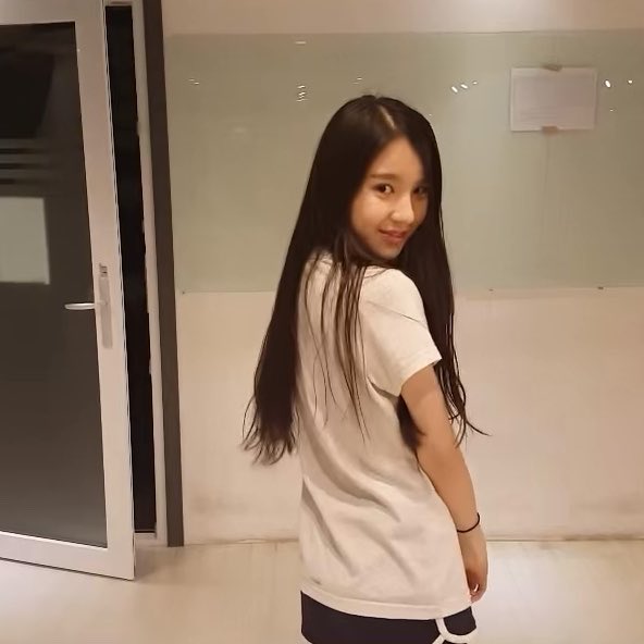 A young woman with long dark hair, wearing a white t-shirt, smiles over her shoulder indoors.